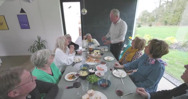 This slow-motion video captures a warm and intimate gathering of senior friends around a dining table in a modern home. The scene shows a cheerful man standing, addressing his friends as they enjoy a - Footage, Video