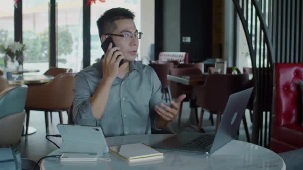 Plan moyen de gestionnaire masculin asiatique occupé parlant avec le client sur un téléphone mobile assis à table devant un ordinateur portable dans un café - Séquence, vidéo