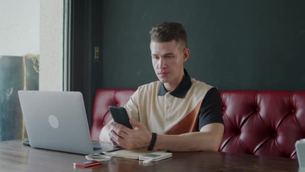 Střední portrét mladého bělocha investora pomocí smartphonu při práci na notebooku v kavárně a při pohledu do kamery - Záběry, video