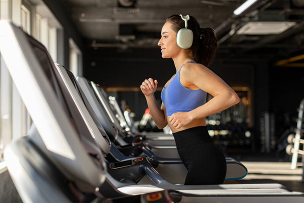 Szczęśliwa wysportowana dama nosząca bezprzewodowe słuchawki podczas joggingu na torze biegowym w siłowni sportowej, ciesząca się treningiem z muzyką - Zdjęcie, obraz