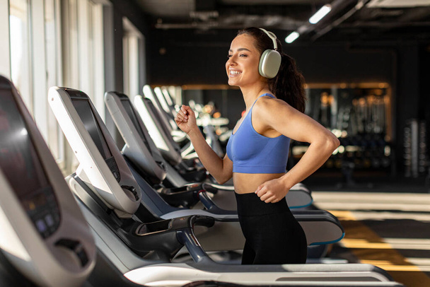Young strong sports woman working out and running on treadmill in gym, listening music in headphones and smiling, enjoying morning workout - Photo, Image