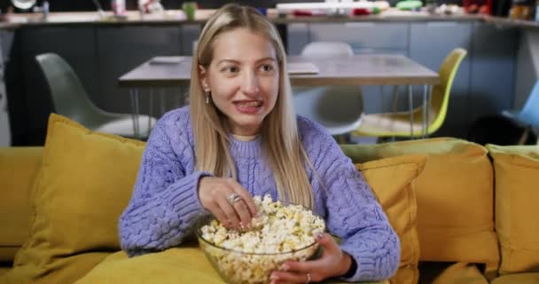 Mujer encantada saborea palomitas de maíz y disfruta de películas mientras está sentado en el sofá en la cómoda sala de estar. Encantadora mujer ríe viendo comedia - Imágenes, Vídeo