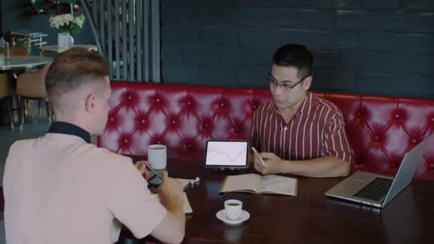 Plan moyen de jeune commerçant caucasien demandant expert asiatique masculin sur les actions prévision pointant vers l'écran de tablette numérique à la table dans le café - Séquence, vidéo
