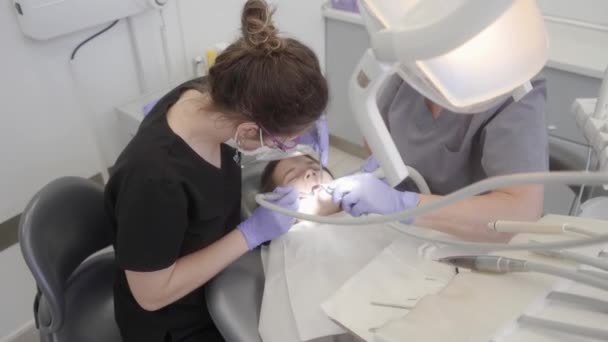 Behandeling van cariës bij een kind in de pediatrische tandheelkunde. De tandarts boort de tandglazuur met een boor, de assistent helpt de arts om een medische procedure uit te voeren. - Video