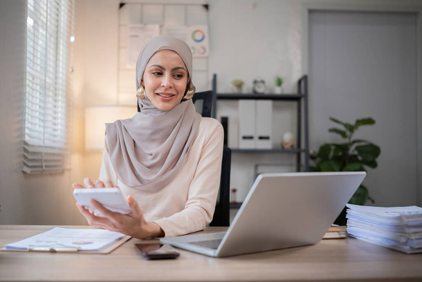 Jovem mulher de negócios muçulmana realiza uma reunião pelo telefone e calcula as finanças da empresa em sua mesa de trabalho.. - Foto, Imagem