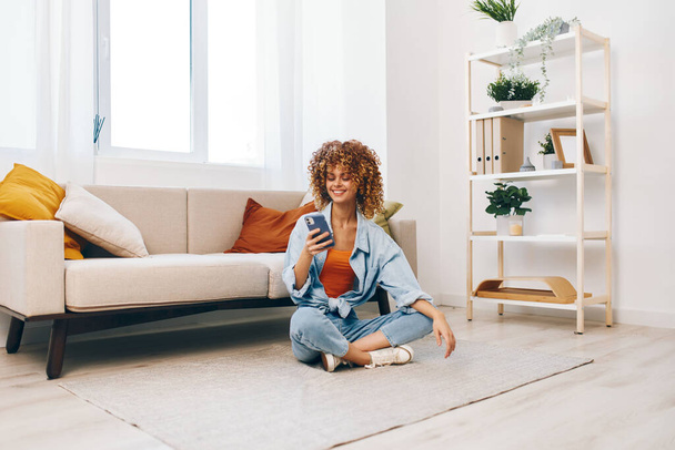 Acogedora comodidad en el hogar: Mujer joven sonriendo y sosteniendo el teléfono móvil, relajarse en el sofá en la sala de estar, disfrutar de juegos en línea y chat - Foto, Imagen