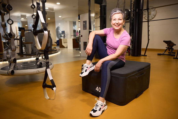 Mature smiling caucasian woman wearing sportswear sitting and resting on sport box and looking at camera during break in gym. Concept of modern healthy lifestyle - Photo, Image