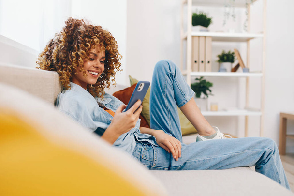 Mulher feliz sentada no sofá, segurando seu telefone celular e sorrindo enquanto joga jogos online - Foto, Imagem