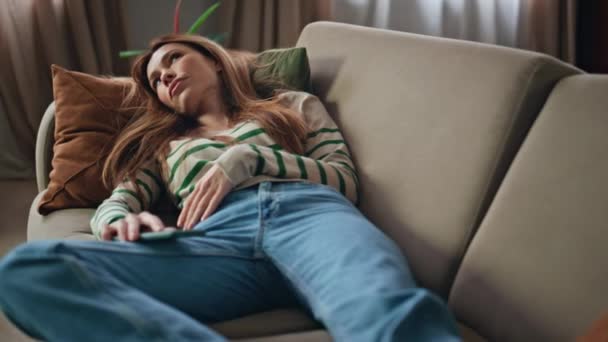 Egy alvó nő a kanapén tartja a mobilját. Vonzó fiatal lány fekszik kényelmes kanapé érzés unatkozik otthon hétvégén. Mosolygós hölgy pihenés egyedül után kemény nap gondolkodás probléma. - Felvétel, videó