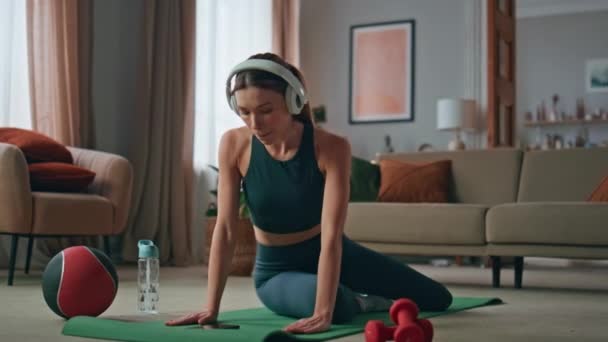 Фітнес-дівчина, що практикує дошку, слухає музику в навушниках вдома. Орієнтована спортивна жінка має ранкове тренування на спортивному килимку в квартирі. Здорова струнка леді в навушниках тренування м'язів тіла в будинку. - Кадри, відео