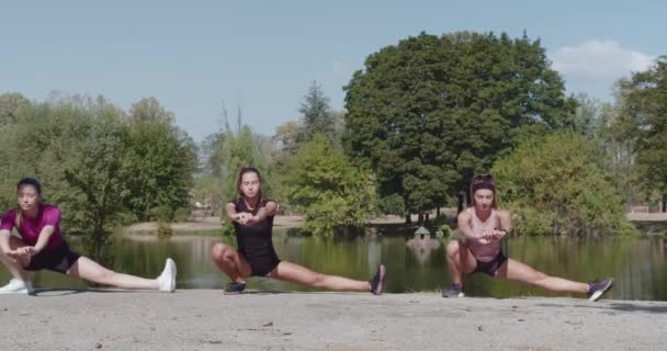Tři fitness nadšenci provádět strečink cvičení podél klidné jezera, podporovat zdraví a wellness v přirozeném prostředí. - Záběry, video