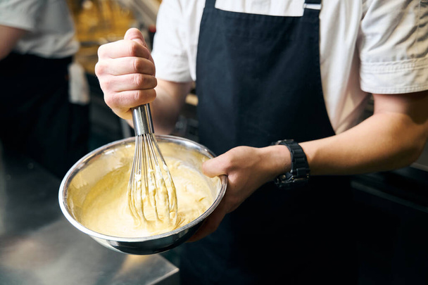 Обрезанный мужчина шеф-повар смешивает тесто с венчиком в миске для приготовления блюд в ресторане. Концепция вкусного здорового питания - Фото, изображение