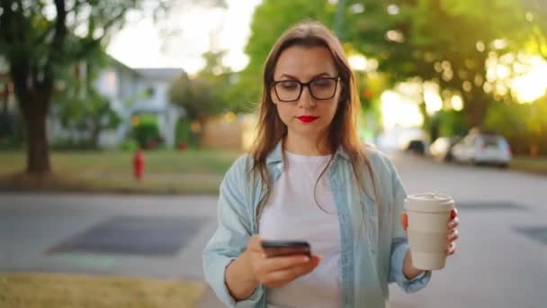 Mujer feliz con el teléfono inteligente y el café en sus manos caminando por el barrio en la noche de verano.  - Imágenes, Vídeo