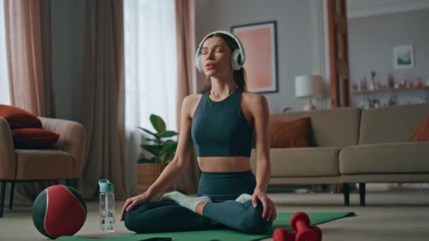 Evde kulaklık takan, Lotus pozisyonunda oturan ilham verici bir kadın. Sakin, kapalı gözlü kız oturma odasında spor minderinde yoga yapıyor. Kablosuz kulaklıkla ses meditasyonunu dinleyen sakin kadın yogi - Video, Çekim