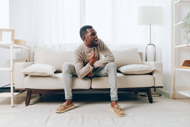 Triste hombre afroamericano sentado en un cómodo sofá en su moderna sala de estar, profundamente pensado Con una leve sonrisa en su rostro, encuentra consuelo en la tranquilidad de su hogar solo con su - Foto, Imagen