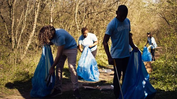 Διαφορετικοί άνδρες εθελοντές μαζεύουν σκουπίδια και πλαστικά σκουπίδια με λαβίδες, εργάζονται για την καταπολέμηση της παράνομης απόρριψης και τη διατήρηση του φυσικού περιβάλλοντος των δασών. Ακτιβιστές εθελοντές για καθαρισμό απορριμμάτων. Κάμερα Β. - Φωτογραφία, εικόνα