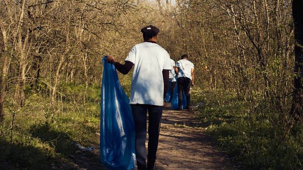 Μια Αφρο-Αμερικανίδα μαζεύει σκουπίδια σε μια σακούλα σκουπιδιών χρησιμοποιώντας λαβίδες, ανακυκλώνοντας πλαστικά απόβλητα και μαζεύοντας σκουπίδια σε ένα δάσος. Μια νεαρή ακτιβίστρια καθαρίζει το δάσος από σκουπίδια. Κάμερα Β. - Φωτογραφία, εικόνα