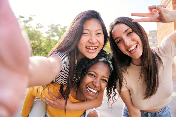Grupo joven multi-étnica hembra tomando retrato sonriente selfie juntos al aire libre. Mujer africana dando paseo a cuestas a la chica asiática en la calle. Emocionados buenos amigos posando para la foto divertida un día soleado - Foto, imagen