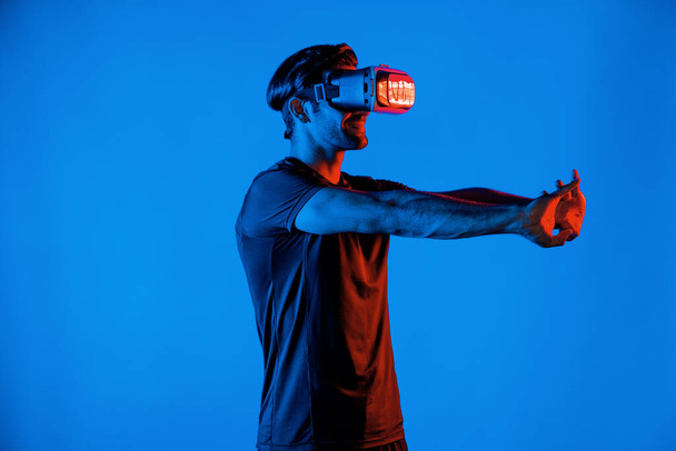Ευτυχισμένος άνθρωπος με VR goggle άσκηση και τέντωμα βραχίονα στο φως νέον φόντο. Καυκάσιος με casual ύφασμα που χαλαρώνει και γυμνάζεται χρησιμοποιώντας τεχνολογία προσομοίωσης εικονικής πραγματικότητας. Απόκλιση. - Φωτογραφία, εικόνα