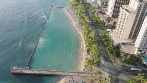 The aerial view showcases Waikiki Beach and the bustling cityscape of Waikiki, Oahu, Hawaii, USA. The video captures the sandy shoreline, turquoise waters, high-rise buildings, and busy streets below. - Footage, Video