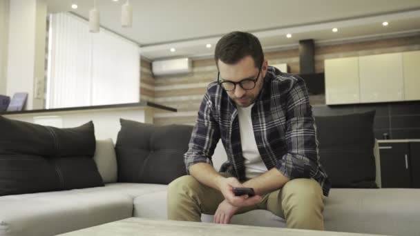 Malheureux jeune homme barbu utilisant smartphone tout en ayant des problèmes, assis à la maison seul. Solitude, dépression, concept émotionnel. Mouvement lent - Séquence, vidéo