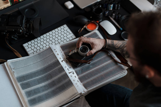 A tattooed photographer carefully examines film negatives at a well-organized desk with photography equipment. - Photo, Image