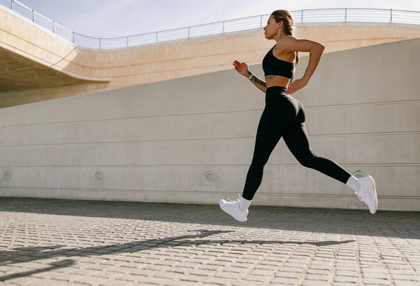 Νεαρή γυναίκα σε αθλητικά ρούχα τρέχει σε σύγχρονα κτίρια φόντο. Έννοια ενεργού τρόπου ζωής - Φωτογραφία, εικόνα