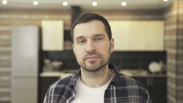 Pozitív fehér szakállas fiatal férfi pózol kockás ingben, miközben otthon ül a konyhában, és a kamerába néz. Életmód koncepció. Lassú mozgás. - Felvétel, videó