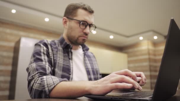 Caucasian bearded young man sitting at the table in kitchen at home and typing on laptop for distance work. Focus on hands. Working online. Slow motion - Footage, Video