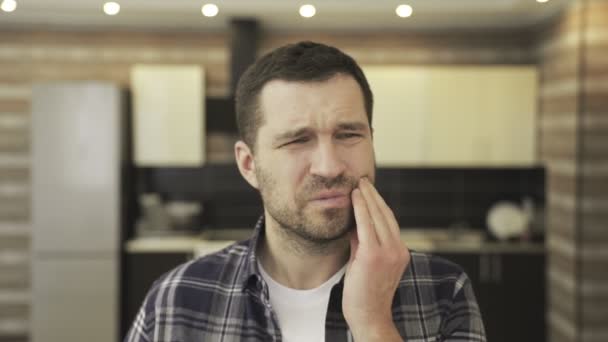Nieszczęśliwy młody brodaty mężczyzna cierpiący na ból zęba w domu podczas siedzenia na kuchni. Ludzie, opieka zdrowotna, stomatologia i koncepcja problemu. Zwolniony ruch - Materiał filmowy, wideo