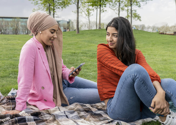 Deux jeunes femmes, l'une portant un hijab, partagent un moment paisible dans un parc, s'engageant avec un smartphone par une journée ensoleillée. - Photo, image