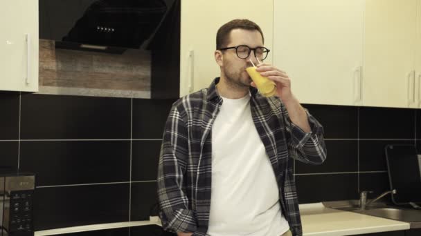 Kaukázusi fickó szemüvegben élvezi a gyümölcslevet, miközben otthon pózol a konyhában. Egészséges táplálkozás, diéta és italok koncepció. Lassú mozgás. - Felvétel, videó