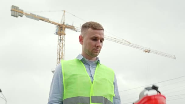 Waist-up portrait of calm builder placing hard hat on his head while standing in front of tower crane. Slow motion - Footage, Video