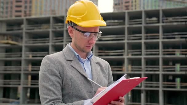 Портрет сфокусованого будівельного аудитора робить нотатки в документах на буфері біля незавершеного багатоповерхового будинку. Повільний рух - Кадри, відео
