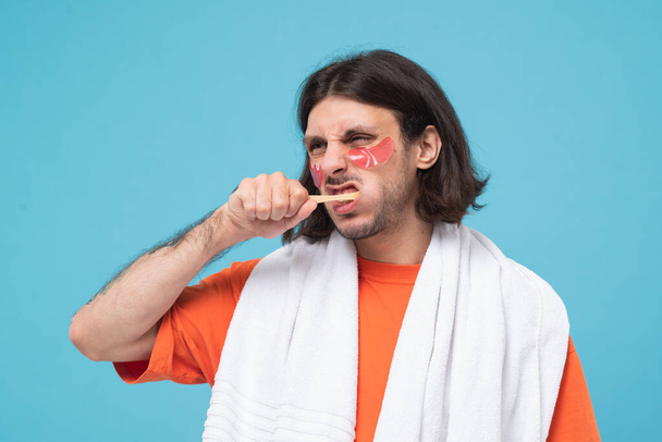 Estúdio tiro de cabelos alegres médio oriental homem escova dentes aplica manchas de hidrogel rosa sob os olhos com toalha branca no fundo azul. - Foto, Imagem