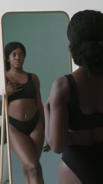 Vertical shot de atractiva mujer negra en sujetador y bragas de pie en el dormitorio en casa y disfrutando de su reflejo en el espejo - Imágenes, Vídeo