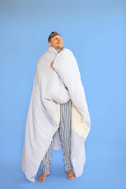 Vol lichaam jongeman in pyjama jam slaap oogmasker verpakt in deken dekbed rust ontspannen thuis geïsoleerd op blauwe achtergrond studio portret. Goede stemming nacht dutje concept. - Foto, afbeelding