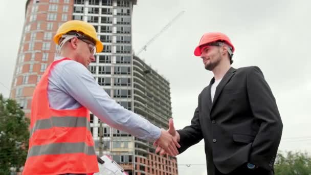 Přátelský inženýr držící půdorys a potřásající si rukou s potěšeným stavebním manažerem poblíž nedokončeného obytného domu. Zpomalený pohyb - Záběry, video