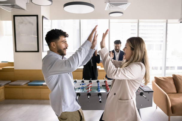 Happy couple of office workers beating colleagues in toy football, giving high five at table soccer stand, smiling, laughing, enjoying win, team success, recreation on work break - Foto, Imagem