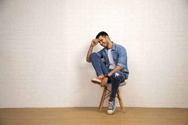 Stylish man in blue jeans and denim shirt is posing while sitting on chair over white background - Foto, Imagem