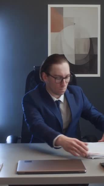 Retrato vertical mediano del oficinista caucásico en traje, gafas, con el pelo liso sentado en el escritorio, alisando la pila de papeles, mirando a la cámara, sonriendo, posando con las manos apretadas - Imágenes, Vídeo