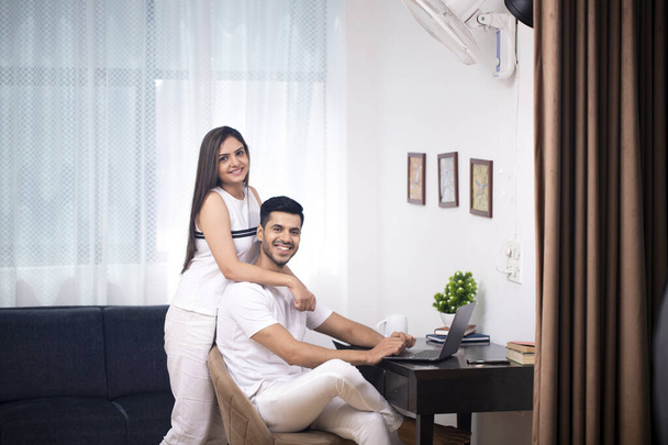 Aziatisch paar in wit outfit zitten in woonkamer en echtgenoot zitten op stoel en vrouw staan achter hem - Foto, afbeelding
