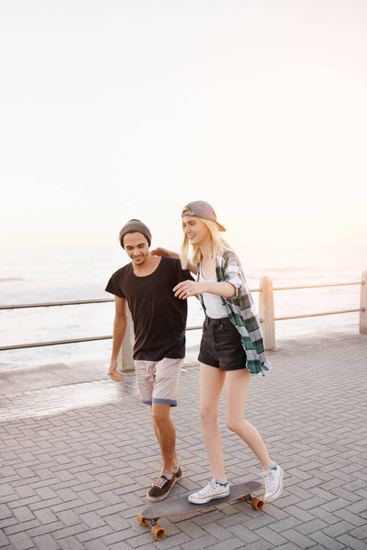 Couple, skateboard and outdoor on boardwalk, happy and helping hand for balance, learning and playful by sea. Man, woman and teaching for skating, support and fun on promenade for vacation in Italy. - Photo, Image