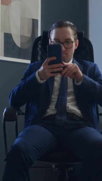 Media verticale di carriera tossica uomo in abito da lavoro, cravatta e occhiali seduto sulla sedia alla scrivania, utilizzando smartphone, inciampando collega a piedi con documenti, indicando il dito, ridendo ferocemente - Filmati, video
