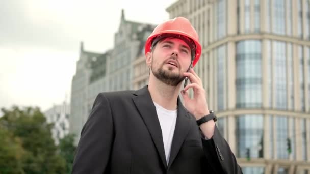 Construtor em terno de escritório e protetora smartphone capacete laranja, chamando e falando. Vídeo em câmera lenta - Filmagem, Vídeo