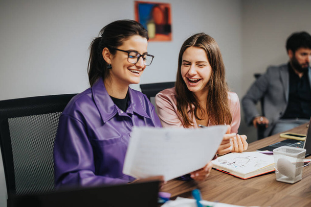 Two young female entrepreneurs laugh while reviewing project charts at a lively startup meeting, showcasing teamwork and productivity in a multicultural office environment. - Photo, Image
