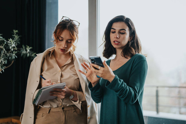 Two professional businesswomen in a discussion, reviewing project specifics, budget, and strategies on a smart phone. - Photo, Image