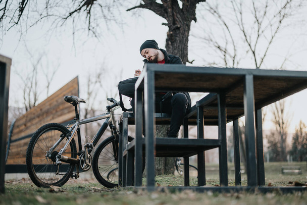 Um jovem senta-se em um banco de parque, tendo uma chamada telefônica, com sua bicicleta de montanha estacionada ao lado dele em um ambiente sereno ao ar livre. - Foto, Imagem