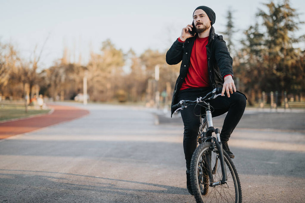 Lässig gekleideter Mann unterbricht seine nachmittägliche Fahrradtour in einem malerischen Park, um zu telefonieren, und zeigt eine Mischung aus Entspannung und Konnektivität. - Foto, Bild
