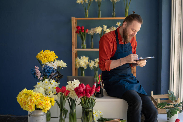 Kaukaski właściciel kwiaciarni nosi fartuch trzymając podkładkę w rękach podczas inwentaryzacji. Przestrzeń kopiowania - Zdjęcie, obraz